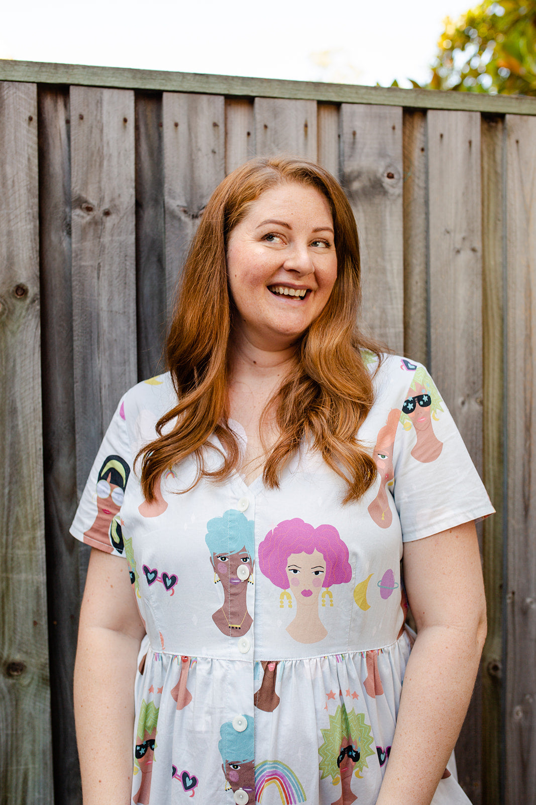 Jane Elliott founder of Brighter Day adaptive clothing wearing a dress with womens faces on it and is standing outside in front of a grey fence. She is smiling and looking up away from the camera