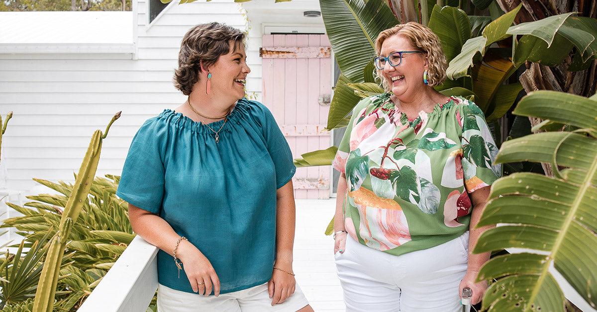 two people wearing adaptive clothing with elastic necklines. They are standing looking at each other and smiling. One woman has a mobility aid