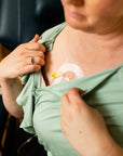 close up of a woman wearing a green adaptive top with the zip open on one side. It shows her chest port IV has been connected and she is receiving chemotherapy for cancer