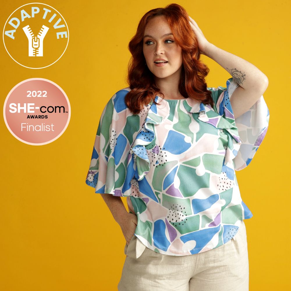 woman wearing an adaptive shirt with a colourful print. she has red hair and looking to the side. Adaptive icon and She-com finalist icon appears on the left