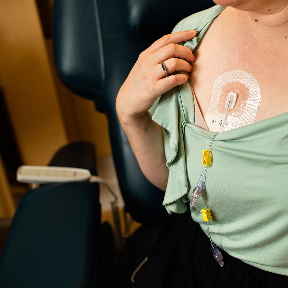 a woman is looking down and has unzipped her green top to reveal a chest port which she is receiving cancer treatment