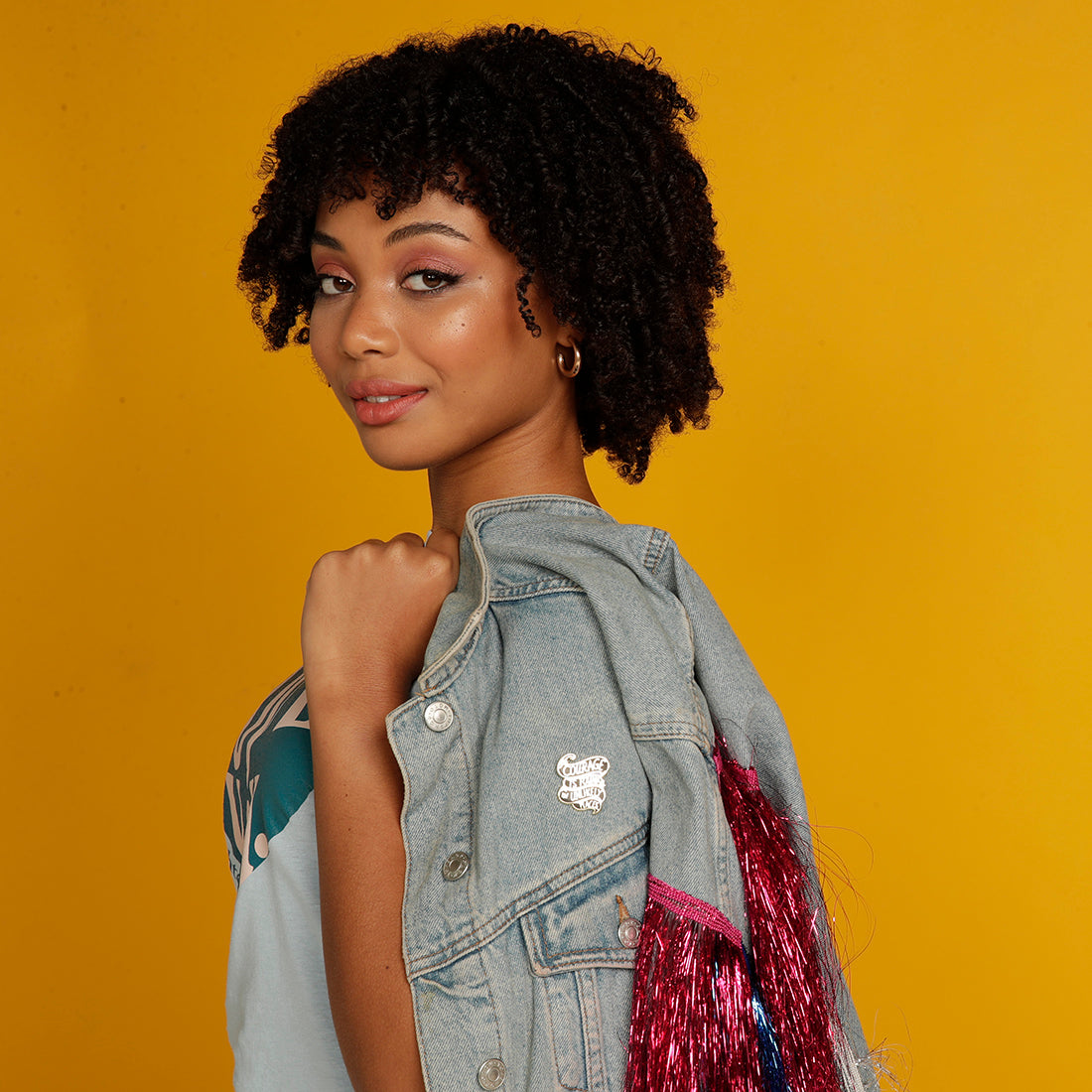 Curly haired woman wearing denim jacket with Courage is found in unlikely places enamel pin