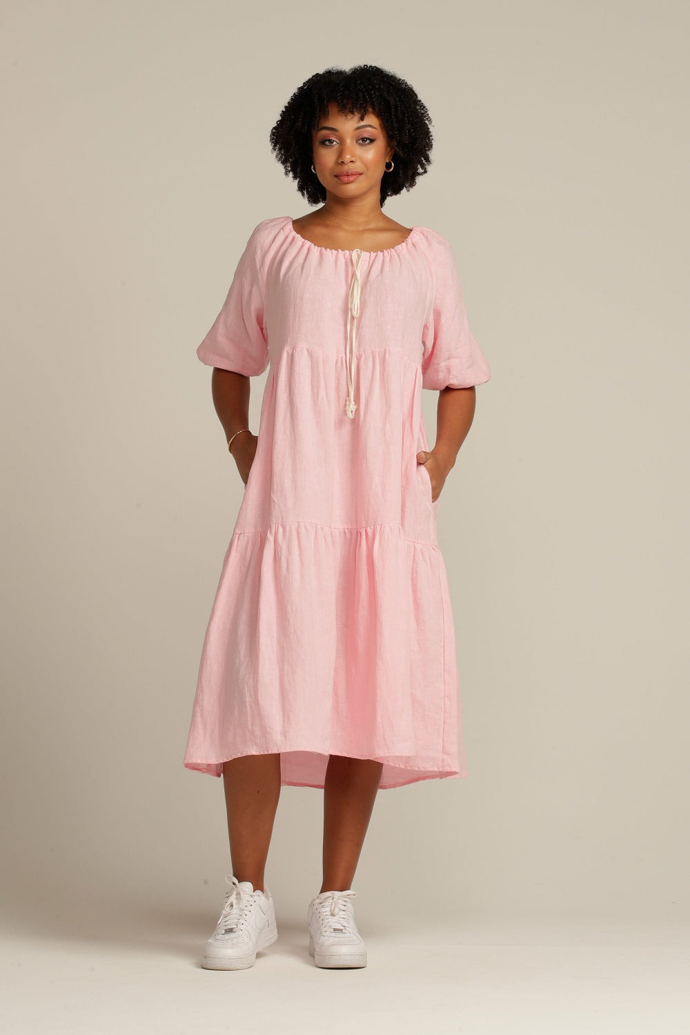 brown woman with black hair is wearing a linen dress that is light pink, she has her hands in the pockets