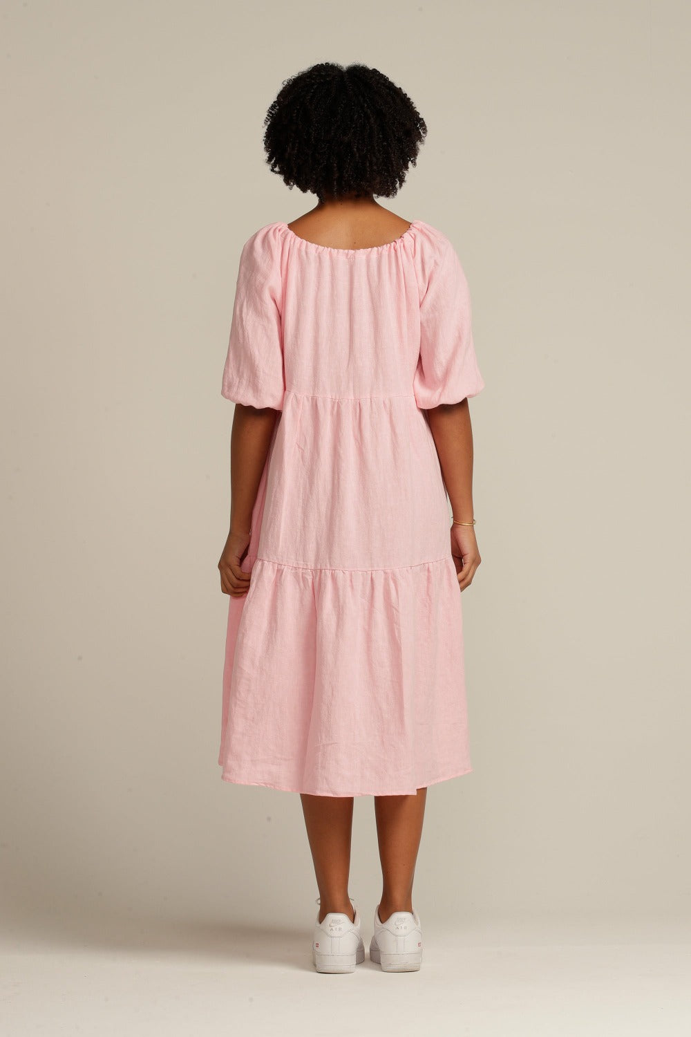 woman with black curly hair facing her back to camera is wearing a pink linen dress that is below knee length